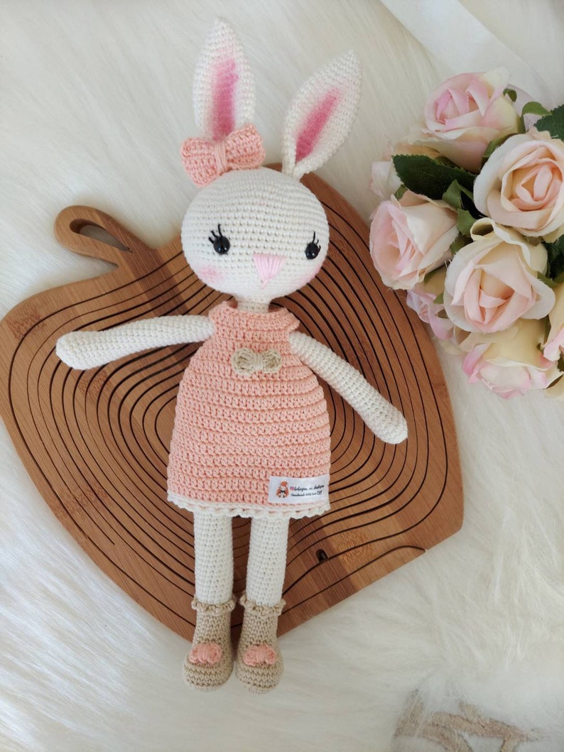 May include: A white crocheted bunny doll wearing a pink dress with a bow. The doll has black eyes and a pink nose. The doll is sitting on a wooden surface.