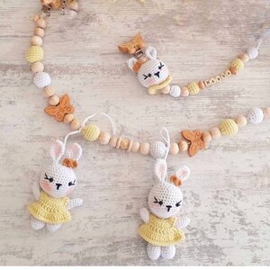 May include: A wooden baby teether with a yellow and white color scheme. The teether features a bunny, butterfly, and wooden beads. The teether is personalized with the name "HANYA".