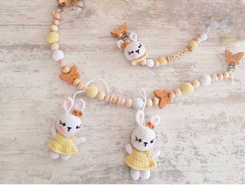 May include: A wooden baby teether with a yellow and white color scheme. The teether features a bunny, butterfly, and wooden beads. The teether is personalized with the name "HANYA".