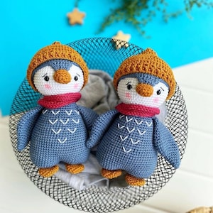 May include: Two crocheted penguin toys with brown hats, red scarves, and blue bodies. The penguins are sitting in a black wire basket.