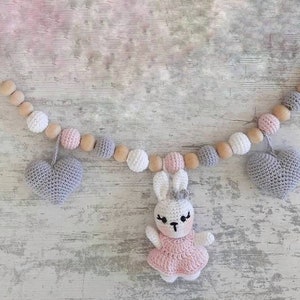 May include: A baby mobile with wooden beads, crocheted hearts, and a crocheted bunny wearing a pink dress. The mobile is hanging from a wooden clip.