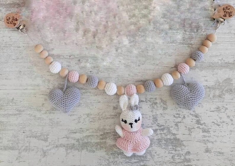 May include: A baby mobile with wooden beads, crocheted hearts, and a crocheted bunny wearing a pink dress. The mobile is hanging from a wooden clip.