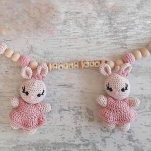 May include: A pink and white crocheted bunny baby toy with a wooden bead and clip chain. The chain has wooden beads with the name "Hafsa Nur" spelled out in wooden letters.