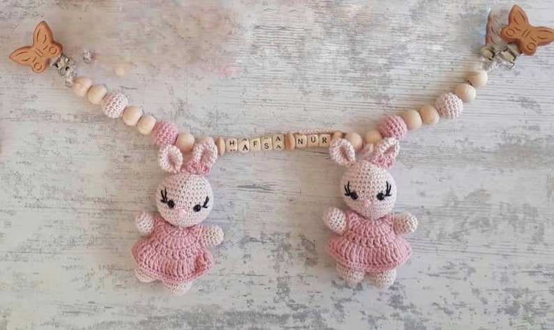 May include: A pink and white crocheted bunny baby toy with a wooden bead and clip chain. The chain has wooden beads with the name "Hafsa Nur" spelled out in wooden letters.