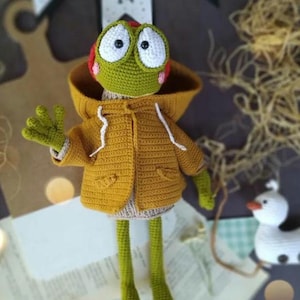 May include: A handmade crochet frog doll wearing a mustard yellow hooded jacket. The frog has large white eyes, green skin, and red accents. The doll is standing on a book with a small white duck figurine nearby.