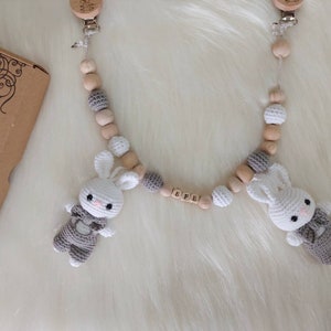 May include: A wooden baby teether with two crocheted bunny toys attached. The teether has wooden beads and a gray and white crocheted bead. The bunny toys have gray and white crocheted bodies and white faces. The teether has a wooden name tag with the name "BREE" on it.