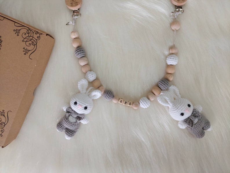 May include: A wooden baby teether with two crocheted bunny toys attached. The teether has wooden beads and a gray and white crocheted bead. The bunny toys have gray and white crocheted bodies and white faces. The teether has a wooden name tag with the name "BREE" on it.