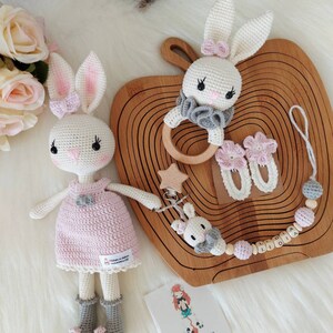 May include: Two crocheted bunny toys, one wearing a pink dress and the other a gray ruffle collar. The bunnies are on a wooden tray with a wooden ring, two crocheted flower clips, and a wooden bead necklace with the name "LIDIA" spelled out.