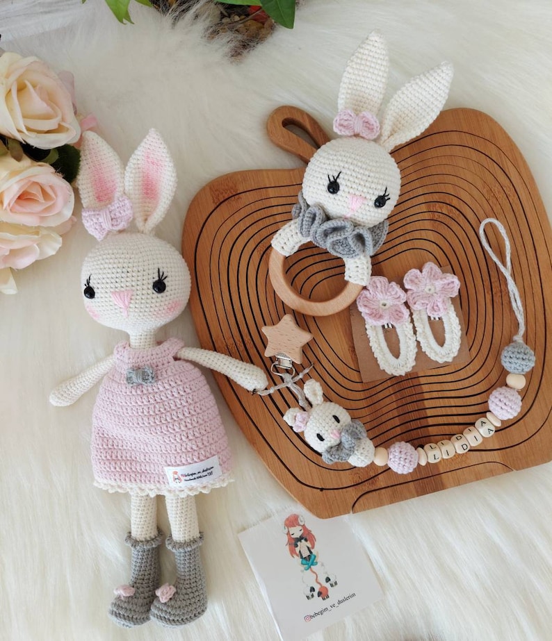 May include: Two crocheted bunny toys, one wearing a pink dress and the other a gray ruffle collar. The bunnies are on a wooden tray with a wooden ring, two crocheted flower clips, and a wooden bead necklace with the name "LIDIA" spelled out.