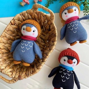May include: Three crocheted penguin toys with different colored hats and scarves. The penguins are sitting on a white surface and in a wicker basket.