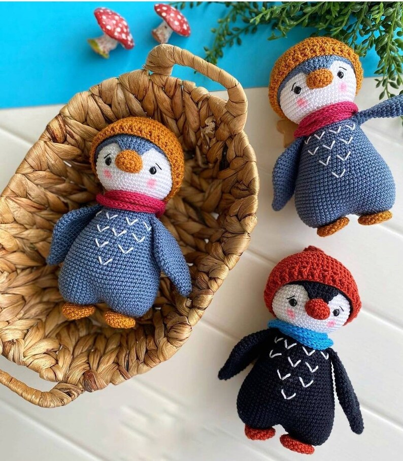 May include: Three crocheted penguin toys with different colored hats and scarves. The penguins are sitting on a white surface and in a wicker basket.