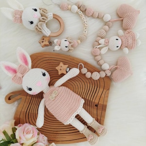 May include: A set of crocheted bunny toys with pink and white accents. The set includes a large bunny with a pink dress, a smaller bunny with a pink dress, a heart-shaped toy, and a wooden bead necklace with the name "NATALIA" spelled out.