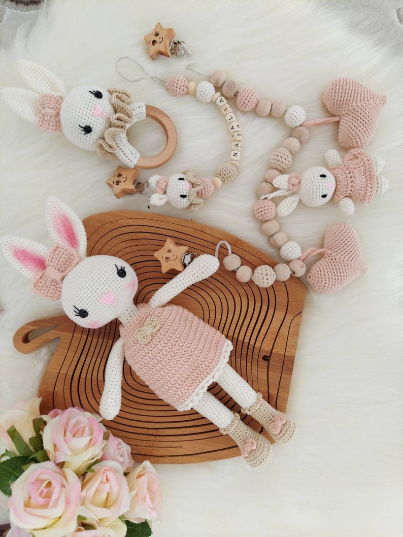 May include: A set of crocheted bunny toys with pink and white accents. The set includes a large bunny with a pink dress, a smaller bunny with a pink dress, a heart-shaped toy, and a wooden bead necklace with the name "NATALIA" spelled out.