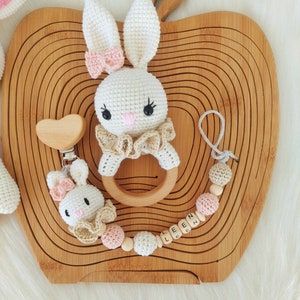 May include: A white crocheted bunny rattle with a wooden ring and a pink bow. A wooden heart-shaped teether is attached to a wooden bead and crocheted bead chain with the name "LEEN" spelled out on wooden beads.