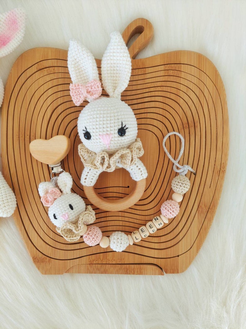 May include: A white crocheted bunny rattle with a wooden ring and a pink bow. A wooden heart-shaped teether is attached to a wooden bead and crocheted bead chain with the name "LEEN" spelled out on wooden beads.