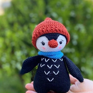 May include: A crocheted penguin toy wearing a red hat and a blue scarf. The penguin has a white face with black eyes and a red nose. The penguin is holding a small white heart in its right hand.