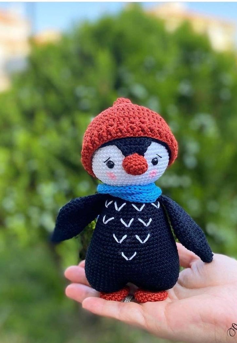 May include: A crocheted penguin toy wearing a red hat and a blue scarf. The penguin has a white face with black eyes and a red nose. The penguin is holding a small white heart in its right hand.