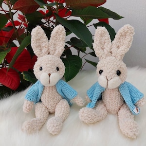May include: Two plush beige bunny rabbits wearing light blue knitted cardigans. The bunnies are sitting on a white fluffy surface.