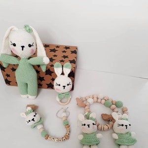 May include: A set of crocheted bunny toys in green and white with wooden beads and a wooden ring. The set includes a large bunny, a smaller bunny with a wooden ring, and a bunny rattle with wooden beads and the name "AH SEN" spelled out.