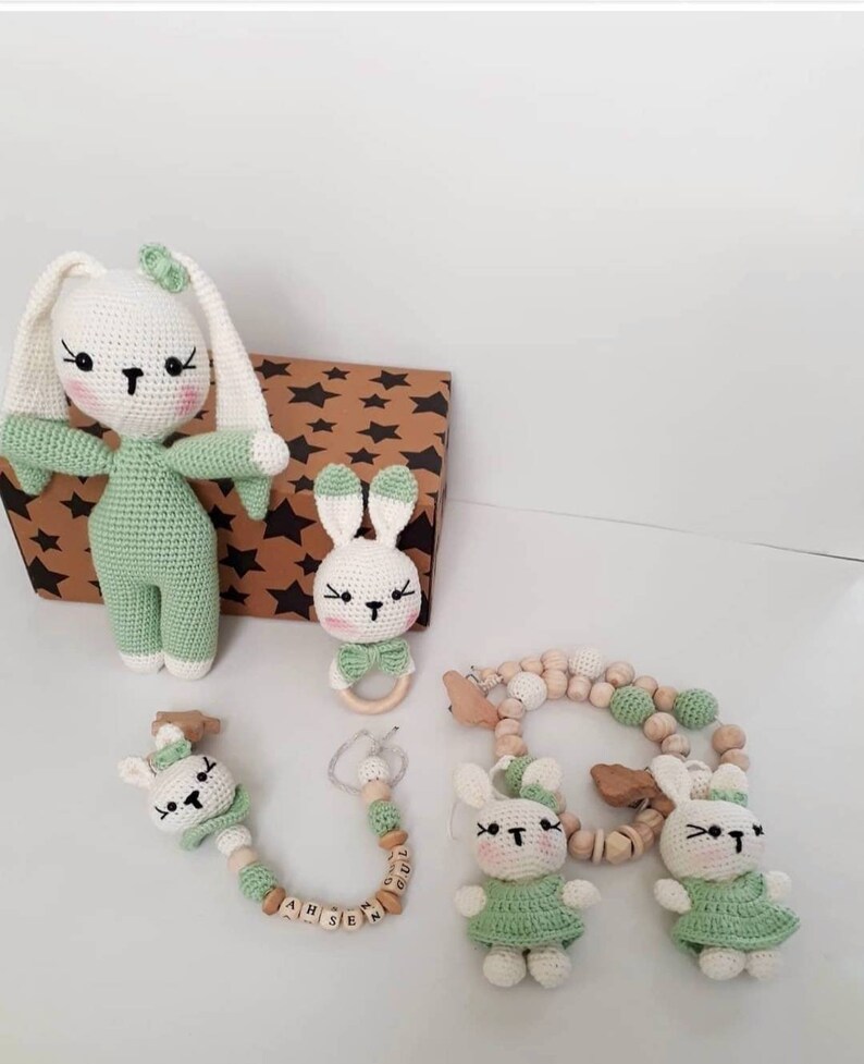 May include: A set of crocheted bunny toys in green and white with wooden beads and a wooden ring. The set includes a large bunny, a smaller bunny with a wooden ring, and a bunny rattle with wooden beads and the name "AH SEN" spelled out.