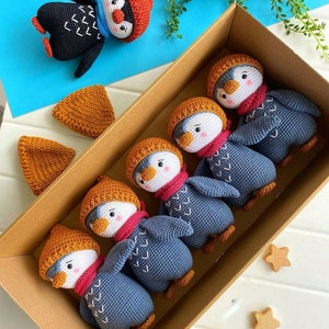 May include: Five crocheted penguin toys in a brown box. The penguins are wearing orange and red hats and scarves. The penguins are blue and gray with white accents. The box is on a white surface with three wooden stars.
