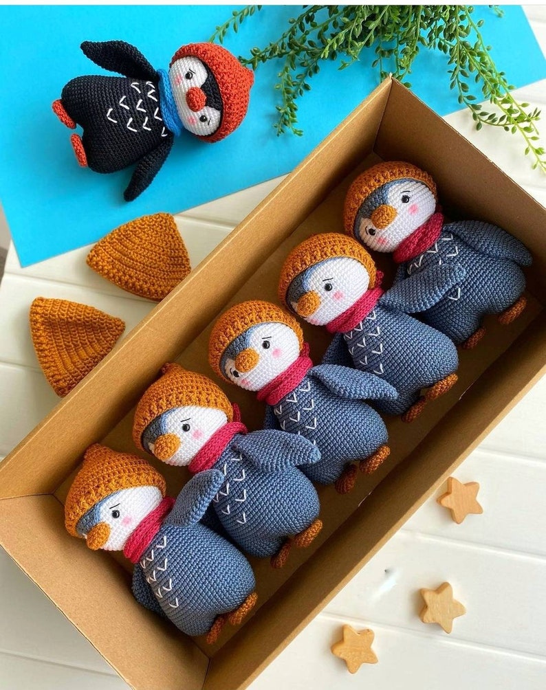 May include: Five crocheted penguin toys in a brown box. The penguins are wearing orange and red hats and scarves. The penguins are blue and gray with white accents. The box is on a white surface with three wooden stars.