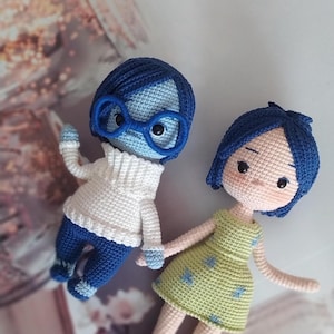 May include: Two crocheted dolls inspired by the characters Sadness and Joy from the movie Inside Out. The doll representing Sadness is wearing a blue outfit and glasses, while the doll representing Joy is wearing a green dress with blue stars.