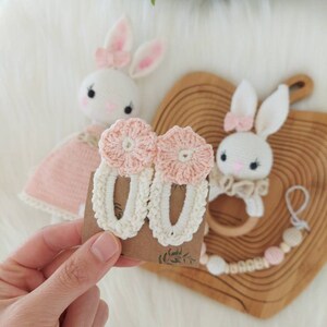 May include: A pair of crocheted hair clips with pink flower accents. The clips are white with a delicate crocheted design.