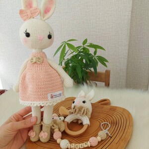 May include: A crocheted pink and white bunny doll with a pink bow, a wooden teething ring, and a wooden bead chain with the name "LEEN" spelled out.