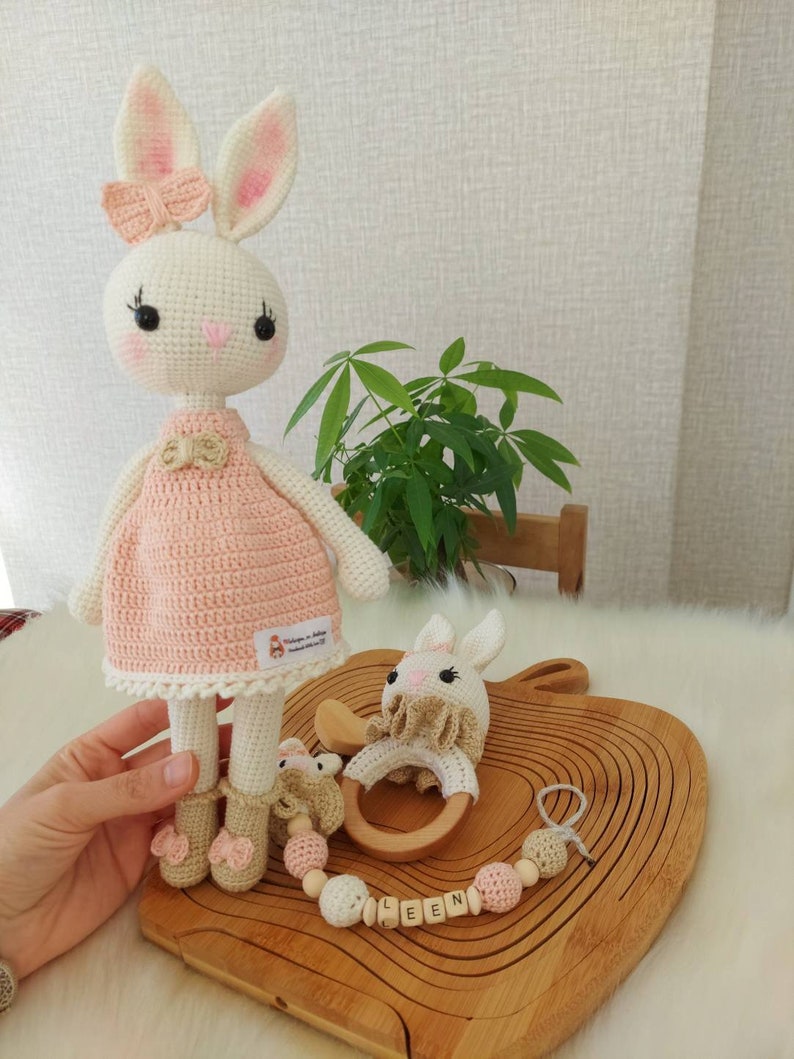 May include: A crocheted pink and white bunny doll with a pink bow, a wooden teething ring, and a wooden bead chain with the name "LEEN" spelled out.