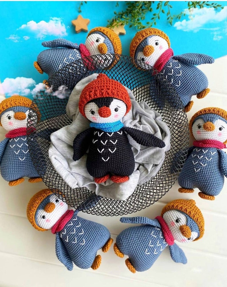 May include: A group of crocheted penguin toys in various colors, including blue, black, and orange. The penguins are wearing hats and scarves and have embroidered details. The toys are arranged in a circle on a black wire mesh surface.