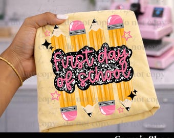 May include: A light yellow t-shirt featuring a graphic print of four yellow pencils with pink erasers. The center displays the text "first day of school" in pink cursive lettering against a black and white speckled background with small stars.