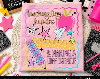 May include: A light pink t-shirt featuring a graphic design with the text "teaching tiny humans & MAKING A DIFFERENCE". The design includes a pink apple, a yellow ruler, a blue and pink checkered pencil, a paper airplane, and various stars.