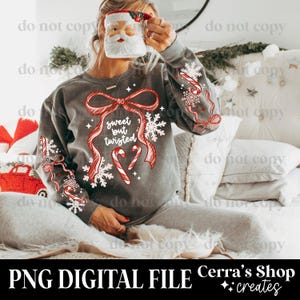 May include: A gray sweatshirt with a festive design featuring a red bow, candy canes, snowflakes, and the text "sweet but twisted." The sleeves have similar decorative elements. A Santa mug is held in the hand.