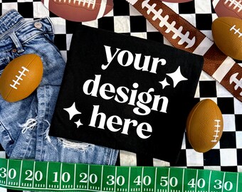 May include: A black t-shirt with the white text your design here and four-pointed star icons, positioned on a black and white checkered background with blue denim jeans, several brown footballs, and a green measuring tape with white numbers.