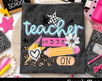 May include: A dark gray t-shirt featuring a graphic design with the text teacher mode on. The design includes a yellow pencil, a white daisy, a black and white checkered star, a heart, and leopard print accents in a vibrant, colorful style.
