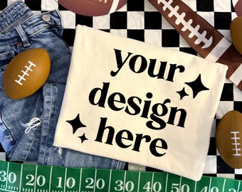 May include: A cream-colored t-shirt featuring the black text 'your design here' and four-pointed star icons. The shirt is displayed alongside blue denim jeans, several brown footballs, and a green measuring tape with white markings.