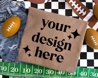 May include: A tan t-shirt with the black text "your design here" and four black star icons. The shirt is surrounded by blue denim jeans, several small brown footballs, and a green measuring tape with white numbers on a black and white checkered background.