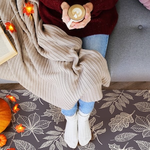 May include: A person sitting on a sofa with a blanket and a book. They are holding a cup of coffee with a leaf design. The sofa is grey and the blanket is beige. The floor is covered in a brown and white rug with a leaf pattern.
