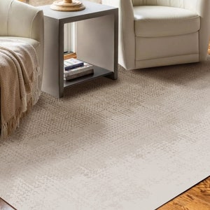 Peut inclure: Un tapis rectangulaire avec un motif beige clair texturé. Le tapis est posé sur un parquet, avec deux fauteuils crème et une table d'appoint visibles en arrière-plan. Une couverture beige est drapée sur l'un des fauteuils.