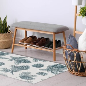 May include: A white and blue patterned doormat with a pine needle design. The doormat is placed in front of a wooden bench with a gray cushion and a wicker basket filled with pillows.