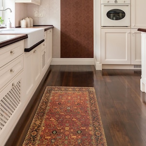 May include: A red, orange and blue patterned rug with a black border in a kitchen with white cabinets and hardwood floors.