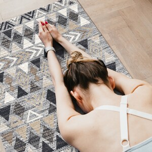 May include: A woman is stretching on a gray and beige yoga mat with a geometric pattern. The mat has a repeating pattern of triangles and arrows.