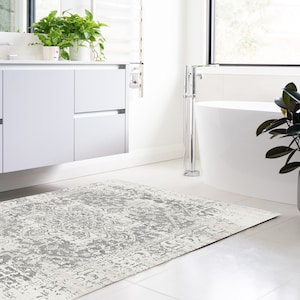 May include: A white bathroom rug with a grey distressed pattern. The rug is placed in front of a white bathtub and a white vanity with a white countertop.