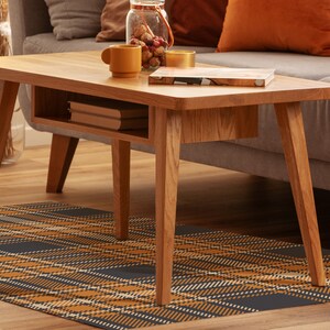 May include: A wooden coffee table with a lower shelf and a plaid rug underneath. The table has a light wood finish and a rectangular shape. The rug is a dark blue and orange plaid pattern.
