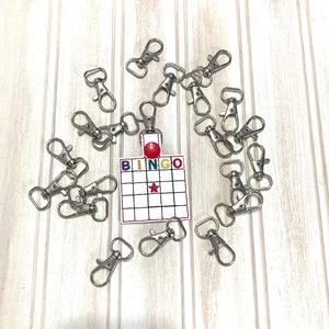 May include: A set of 18 silver keyrings with a white bingo card featuring a red star and a red dot in the centre. The bingo card has the word "BINGO" printed in rainbow colours.
