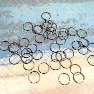 May include: A scattering of small, silver metal rings on a patterned blue and white surface. The rings are circular and made of a shiny metal. They are likely used for jewellery making or crafts. Each ring has an approximate diameter of 1 cm.