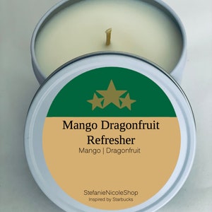 May include: A white candle tin with a green and gold label. The label says "Mango Dragonfruit Refresher" and "Mango | Dragonfruit". The label also says "StefanieNicoleShop Inspired by Starbucks".