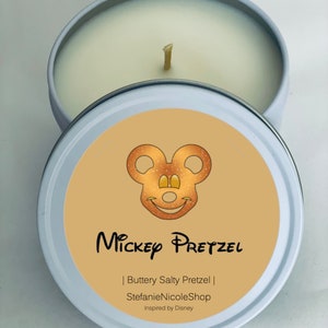May include: A white candle tin with a yellow lid. The lid has a Mickey Mouse design with a pretzel for his head. The text on the lid reads "Mickey Pretzel | Buttery Salty Pretzel | StefanieNicoleShop | Inspired by Disney".