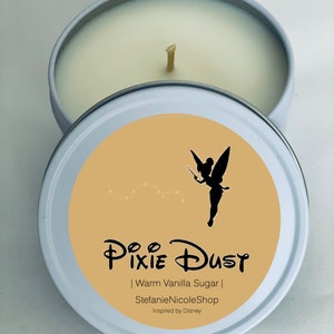 May include: A white candle tin with a beige lid. The lid features a silhouette of Tinkerbell with a wand, and the text "Pixie Dust | Warm Vanilla Sugar | StefanieNicoleShop | Inspired by Disney".